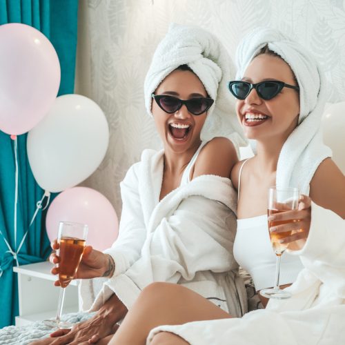 Two young beautiful smiling girls in white bathrobes and towels on head.Sexy carefree women sitting on bed in posh apartment or hotel room.Positive models drinking champagne at bachelorette party